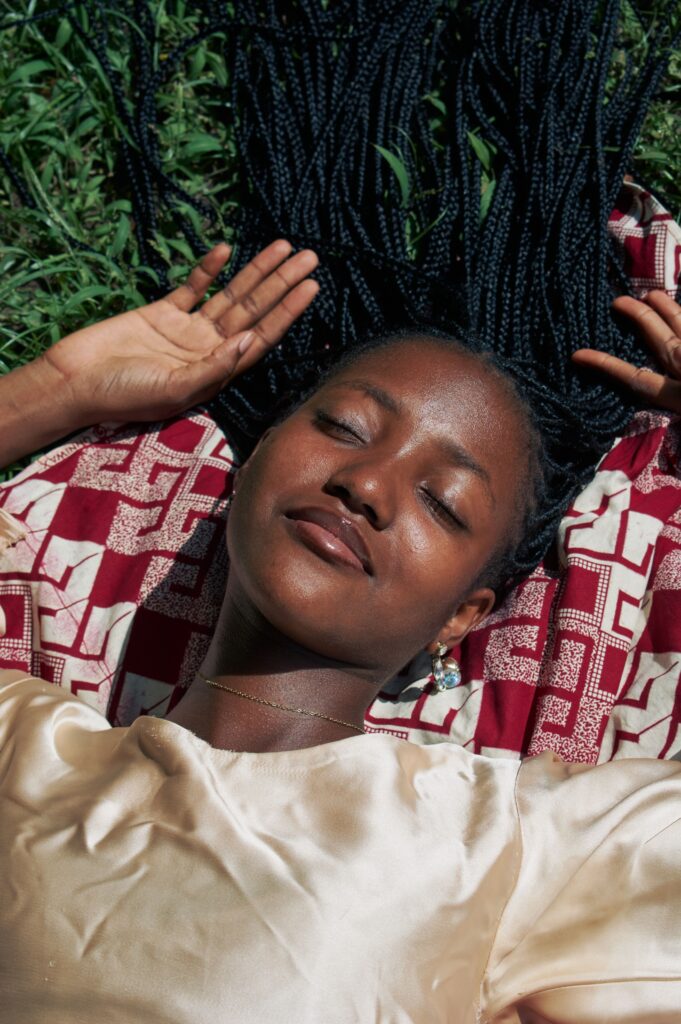 pexels-emmanuel-ijimdiya-1907978092-34031586 A woman with braided hair relaxing on a red patterned blanket in the sun, eyes closed.